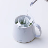 Water being poured from spout of silver teapot into white Ippodo Tea logo mug through Drip Teabag clipped onto top of mug