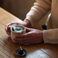 Person in sweater holding mug of steeping tea in both hands with drip teabag clipped onto top beside plate of wagashi dessert
