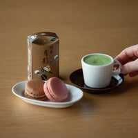 Plate of colorful macarons beside usucha matcha in white espresso cup on brown saucer beside brown decorative box of matcha