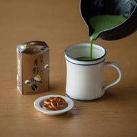 Pouring green prepared matcha from Black Tea Bowl with Spout into mug beside brown floral package and small plate of pretzels