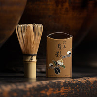 Tan box of Ippodo Tea autumn-exclusive matcha with Japanese flowers and bamboo whisk on rustic shelf among antique tea pots