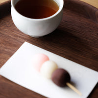 Neopolitan colored wagashi desserts on bamboo skewer set down on white Kaishi Tissue beside cup of steeped tea on wooden tray