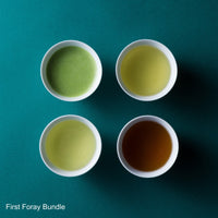 Four white cups with different colored teas on a teal background, labeled 'First Foray Bundle'.