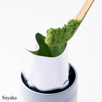 Heaping scoop of green Sayaka matcha powder on chashaku bamboo tea ladle above opened foil pouch in white Ippodo Tea tin