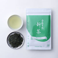 Teacup of light yellow brewed shincha tea beside silver plate of loose rolled tea leaves and green package of New Harvest Tea