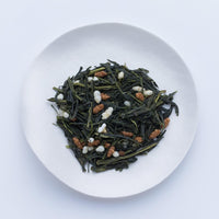 Sample of Ippodo Genmaicha dried bancha green tea leaves and stems mixed with roasted rice on white plate, on white table