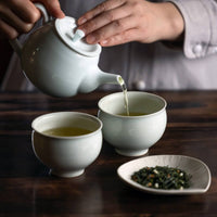 Pouring tea from white porcelain Kyusu into two small porcelain cups beside petal shaped plate of tea leaves on wooden table