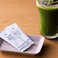 Two Matcha-To-Go Packets on small light purple rectangular plate on wood table beside tall glass of green matcha over ice