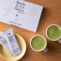 Top view of box of Matcha-To-Go Packets beside two individual packets on yellow saucer and two small mugs of green matcha tea