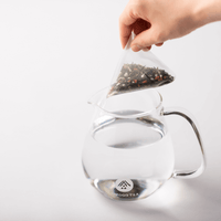 White pyramid-shaped One-Pot Teabag filled with Genmaicha Japanese tea with roasted rice held above glass teapot of water