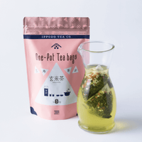 Curvy decorative glass vessel with spout containing two one-pot tea bags in tea beside pink pack of Genmaicha tea with rice