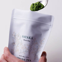 Scooping spoonful of bright green matcha powder out of large white reseleable 100g bag of Sayaka by Ippodo Tea