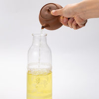 Pouring Ippodo green tea from above from brown Yakishime clay kyusu teapot into glass carafe with lid removed for storing