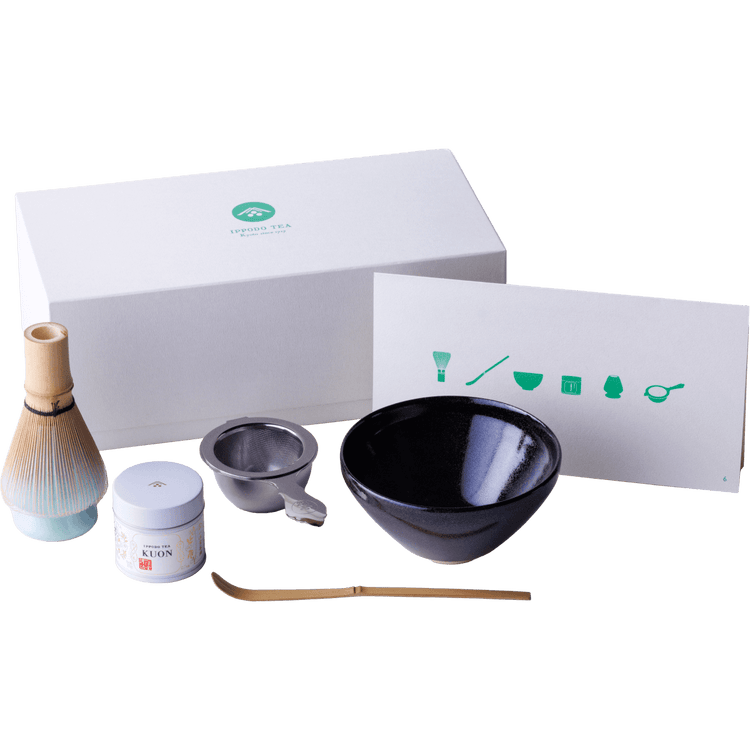 Ippodo gift box beside dark matcha tea bowl, tea strainer in base, Kuon matcha tin, bamboo ladel and whisk on ceramic stand