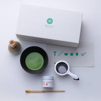Ippodo gift box beside dark matcha tea bowl, tea strainer in base, Kuon matcha tin, bamboo ladel and whisk on ceramic stand