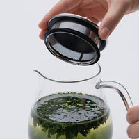 Holding open Ippodo Tea glass teapot containing unfurling looseleaf green tea to display whole-lid circular mesh strainer