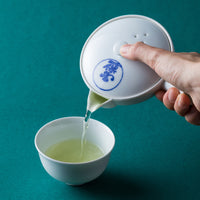 Pouring green tea from white porcelain Kiyomizu-yaki kyusu teapot with blue logo into white porcelain teacup on teal table