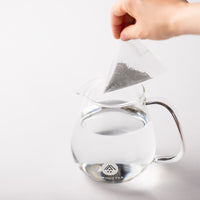 White pyramid-shaped One-Pot Teabag filled with Obukucha Japanese genmaicha green tea dangled over hot water in glass teapot