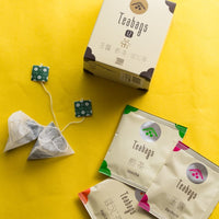 Teabag Assortment box laid on yellow table beside array of different colored individually wrapped teabags and pyramid teabags