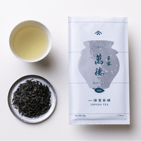 Porcelain teacup of straw colored gyokuro green tea beside plate of dark tea leaves and packaging for Mantoku Ippodo Tea