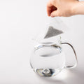 White pyramid-shaped Teapot-Size Teabag filled with Obukucha Japanese genmaicha green tea dangled over water in glass teapot