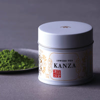 Vivid green matcha powder on artisan silver tin plate beside ornate Japanese can of Ippodo Kanza on grey background