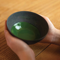 Verdant deep green thick Kanza premium koicha Japanese matcha in dark speckled bowl held with two hands above wooden table