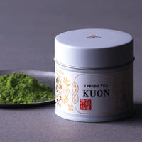 Vivid green matcha powder on artisan silver tin plate beside closed decorative Japanese can of Ippodo Kuon on grey background