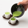 Hand pouring bright green usucha matcha from Black Tea Bowl with Serving Spout into two white espresso cups on brown saucers