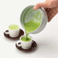 Hand pouring bright green usucha matcha from White Tea Bowl with Serving Spout into two white espresso cups on brown saucers