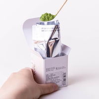 Box of Hatsu matcha with instructions on back and inner foil held open while scooping matcha powder with bamboo tea ladel