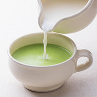 Pouring milk from creamer into light green fresh matcha latte made with Ippodo Tea Ikuyo tea powder in white teacup
