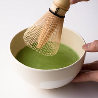 Hand holding cream tea bowl whisking thin usucha Ippodo Tea Wakaki matcha with Chasen bamboo 80-tip whisk white table