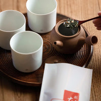 Scooping green tea with rice into clay teapot on lacquered wooden tray with three white minimalist Ippodo-branded teacups