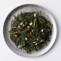 Loose leaf Obukucha Good Fortune Tea with deep green rolled tea and golden rice kernels on silver plate set on white table