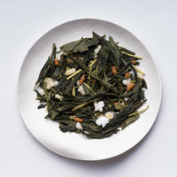 White plate of loose dried dark Japanese green tea blended with roasted rice Organic Genmaicha by Ippodo Tea on white table