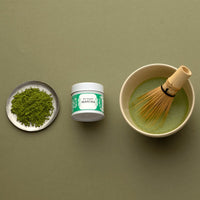 Green matcha tea powder on silver plate beside tin of Organic Matcha and bowl of prepared matcha with whisk on green table