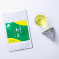 Bright green and yellow packaging for Uji-Shincha laying on white table beside a one-pot teabag and glass of light color tea