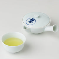 Pouring green tea from white porcelain Kiyomizu-yaki kyusu teapot with blue logo into white porcelain teacup on white table