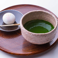 Stone bowl of vivid green thick matcha with Japanese white mochi treat on small plate on lacquered brown wood marubon tray