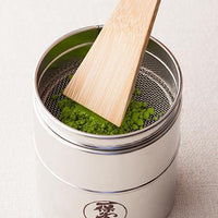 Bamboo takebera spatula sifting bright green matcha in bottom half of tin matcha sieve (burui) with Ippodo Tea logo on front