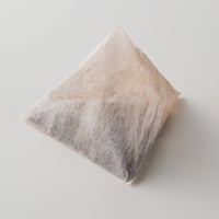 Single pyramid-shaped large one-pot teabag filled with Ippodo Tea Co. mugicha coarsely cracked barley set atop white table