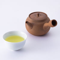 Brown clay Yakishime Kyusu teapot with Ippodo Tea emblem on lid beside white porcelain teacup of light green Japanese tea