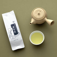 Package of Organic Genmaicha green tea with rice beside ivory clay teapot and white porcelain cup of light tea on green table