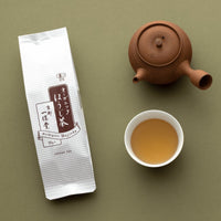 Package of Organic Hojicha roasted green tea beside brown clay teapot and white porcelain cup of amber tea on green table