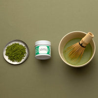 Green matcha tea powder on silver plate beside tin of Organic Matcha and bowl of prepared matcha with whisk on green table