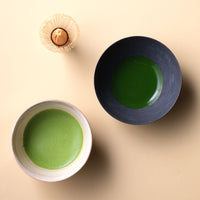 Thin frothy usucha matcha in cream tea bowl and thick koicha matcha in dark tea bowl and chasen bamboo whisk on beige table