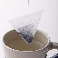 White pyramid shaped teabag filled with Japanese Sencha by Ippodo Tea dangled over beige ceramic mug of hot water