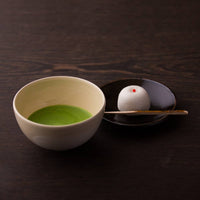 Cream with white brush stroke Mino-yaki Chawan tea bowl containing green matcha beside white Japanese sweet on black plate
