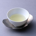 Small white porcelain cup of light color Japanese green tea on silver dual petal leaf shaped saucer on grey table